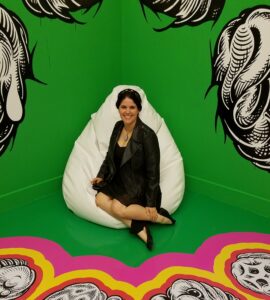 Emma G Rose dressed in black sits on a white beanbag chair in a the corner of a brightly painted art mural