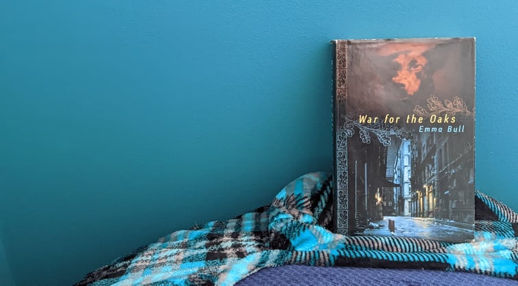 A hardcover edition of War for the Oaks by fantasy author Emma Bull against a blue background with a plaid blanket beneath